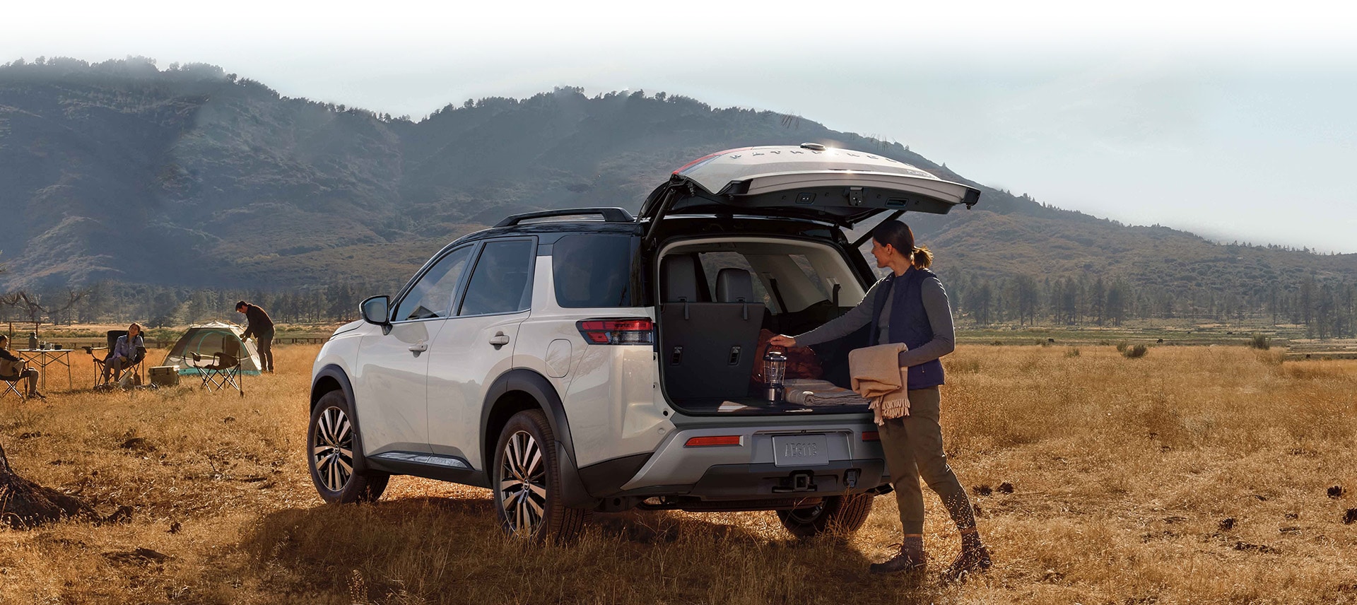 2024 Nissan Pathfinder at a campsite, rear view, with the rear door open and someone getting camping supplies from the cargo area.